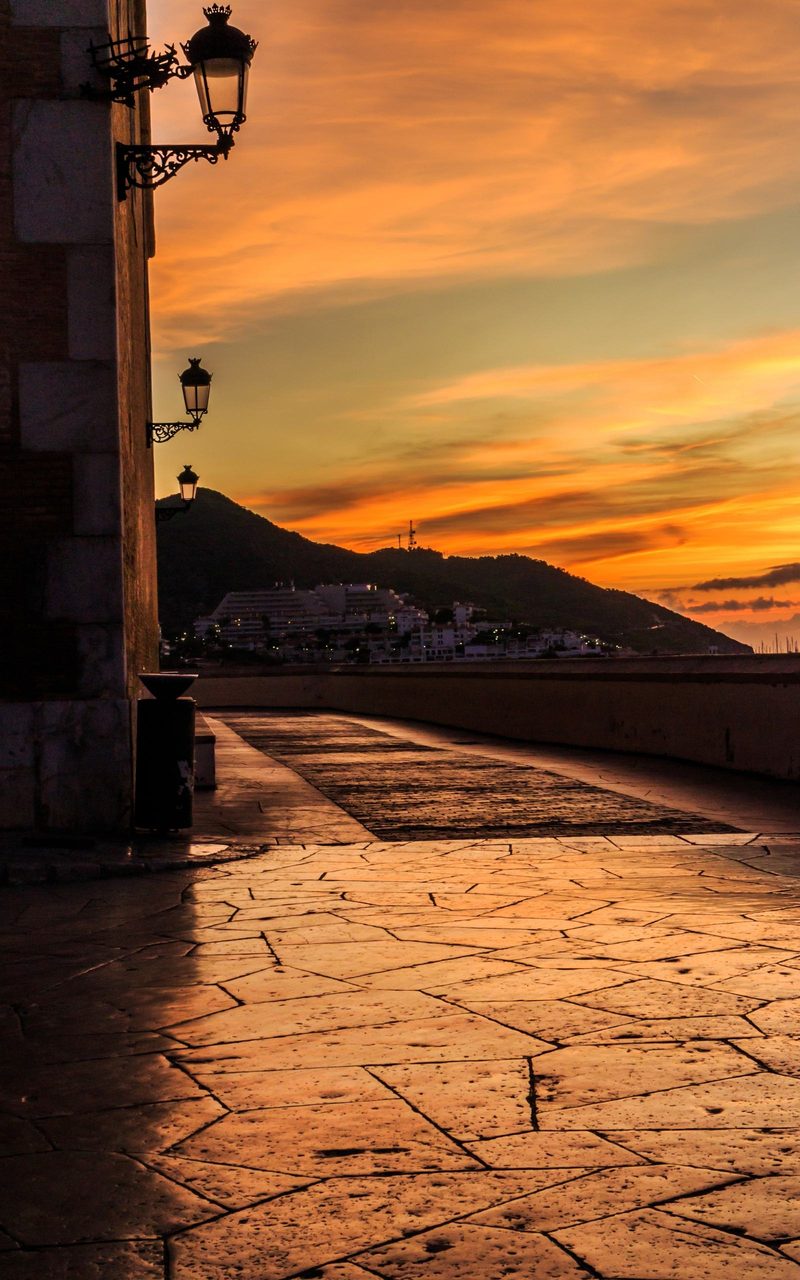 The seafront terrace beside the church of Sant Bartomeu in Sitges at sunset, iron lamps on the promenade