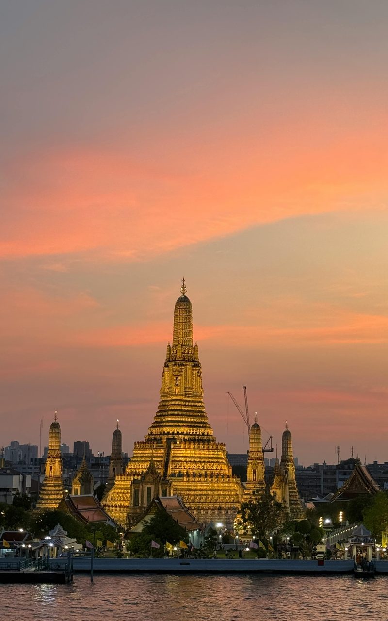 Wat Arun on the Chao Phraya river in Bangkok at sunset, its spires lit gold against a pink sky